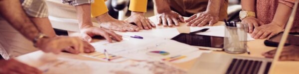 Plusieurs mains de personnes différentes posées sur une table sur laquelle sont disposées des feuilles, une tablette numérique et un ordinateur