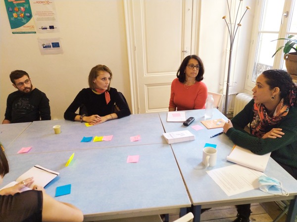 Photo lors d'un atelier collaboratif avec 5 personnes autour d'une table, une personne qui parle et les autres l'écoutent. Des posts its sont sur la table